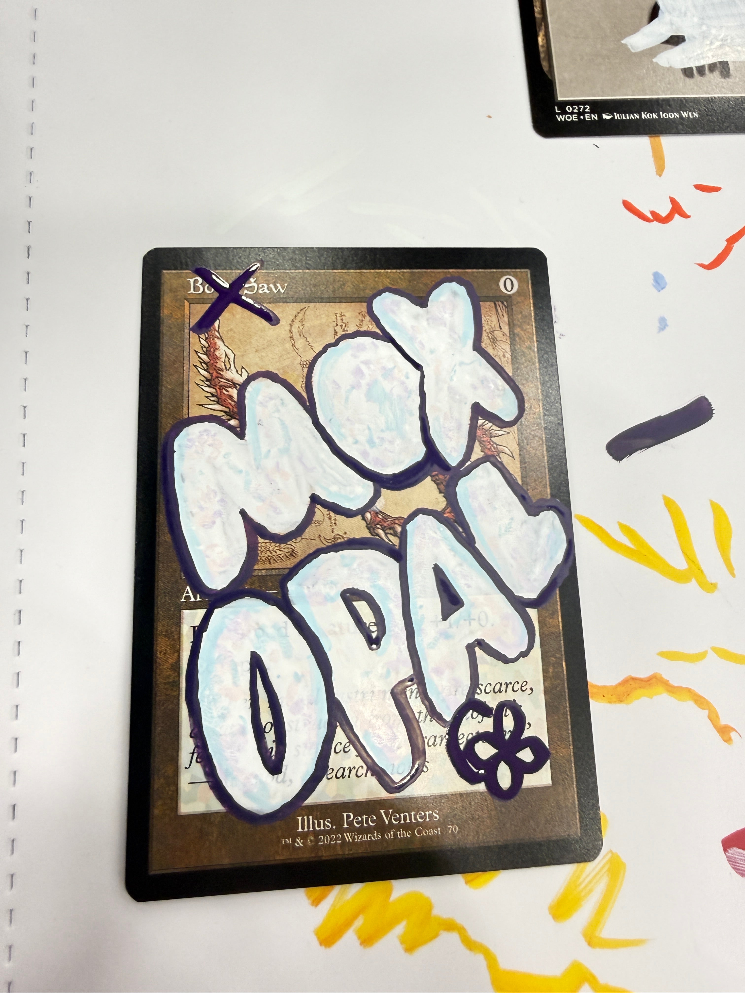 A collectible card featuring a fantasy map design with large white graffiti-style lettering overlaid across the front, photographed on a white surface with colorful marker marks in the background.