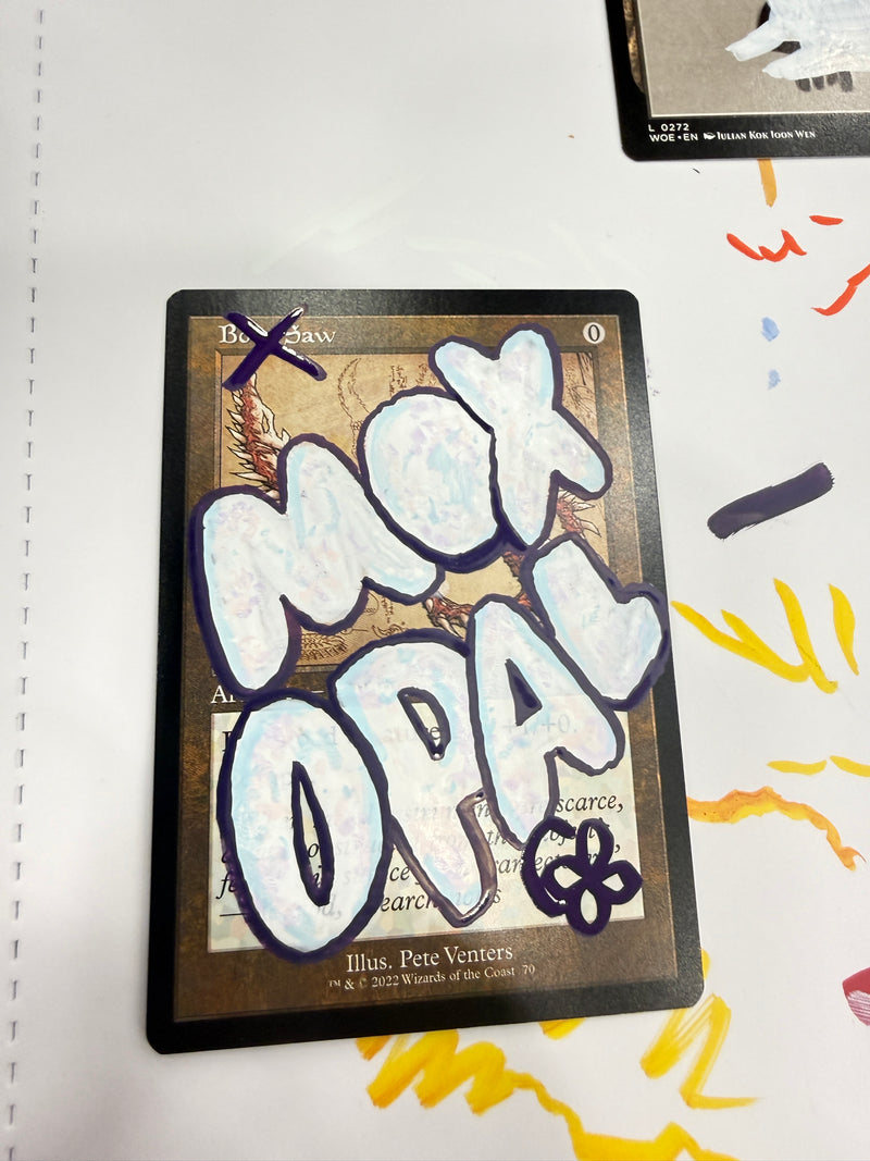 A collectible card featuring a fantasy map design with large white graffiti-style lettering overlaid across the front, photographed on a white surface with colorful marker marks in the background.