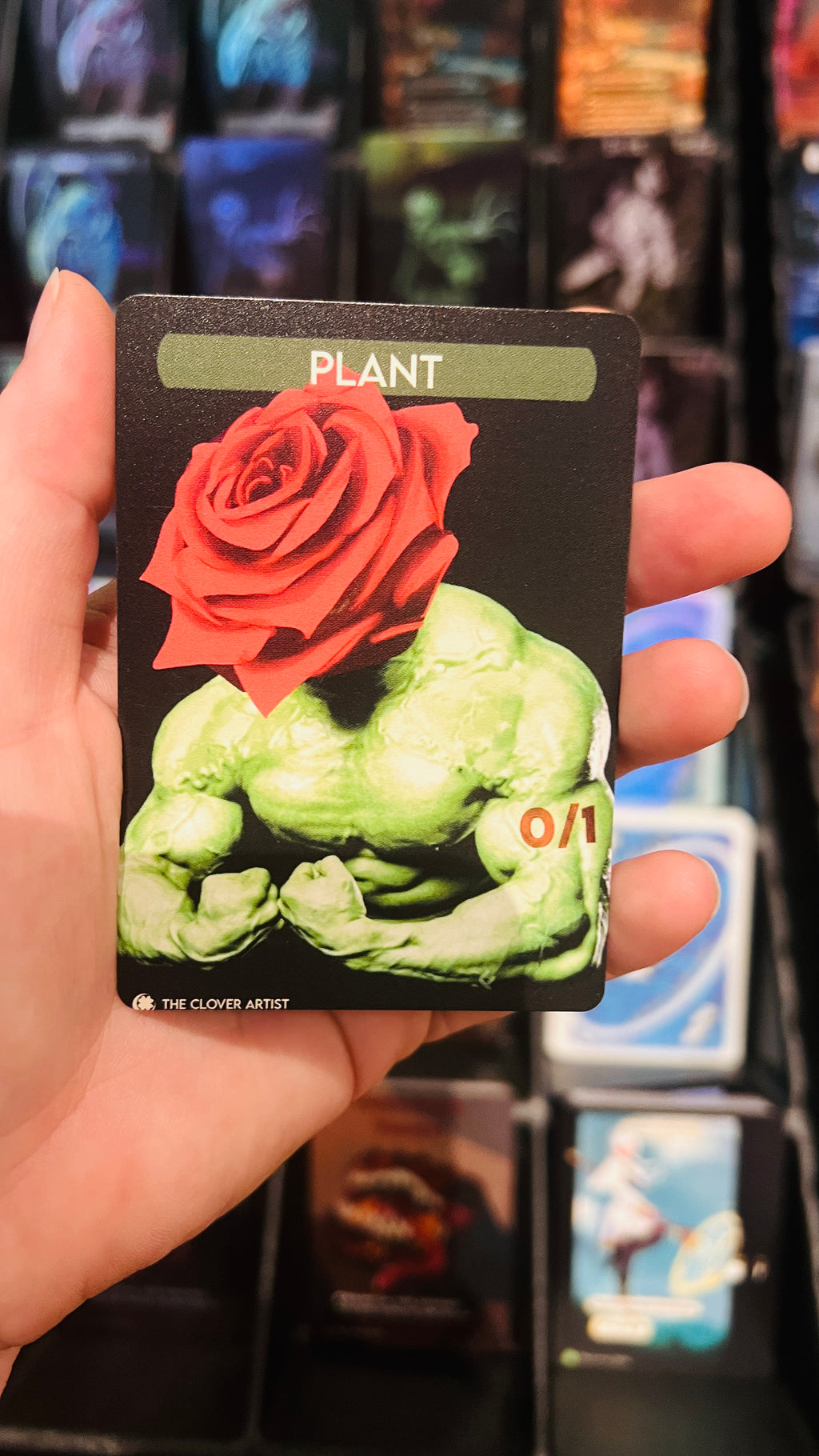 Card with a green muscular figure and red rose held by a hand against a blurred card display background.