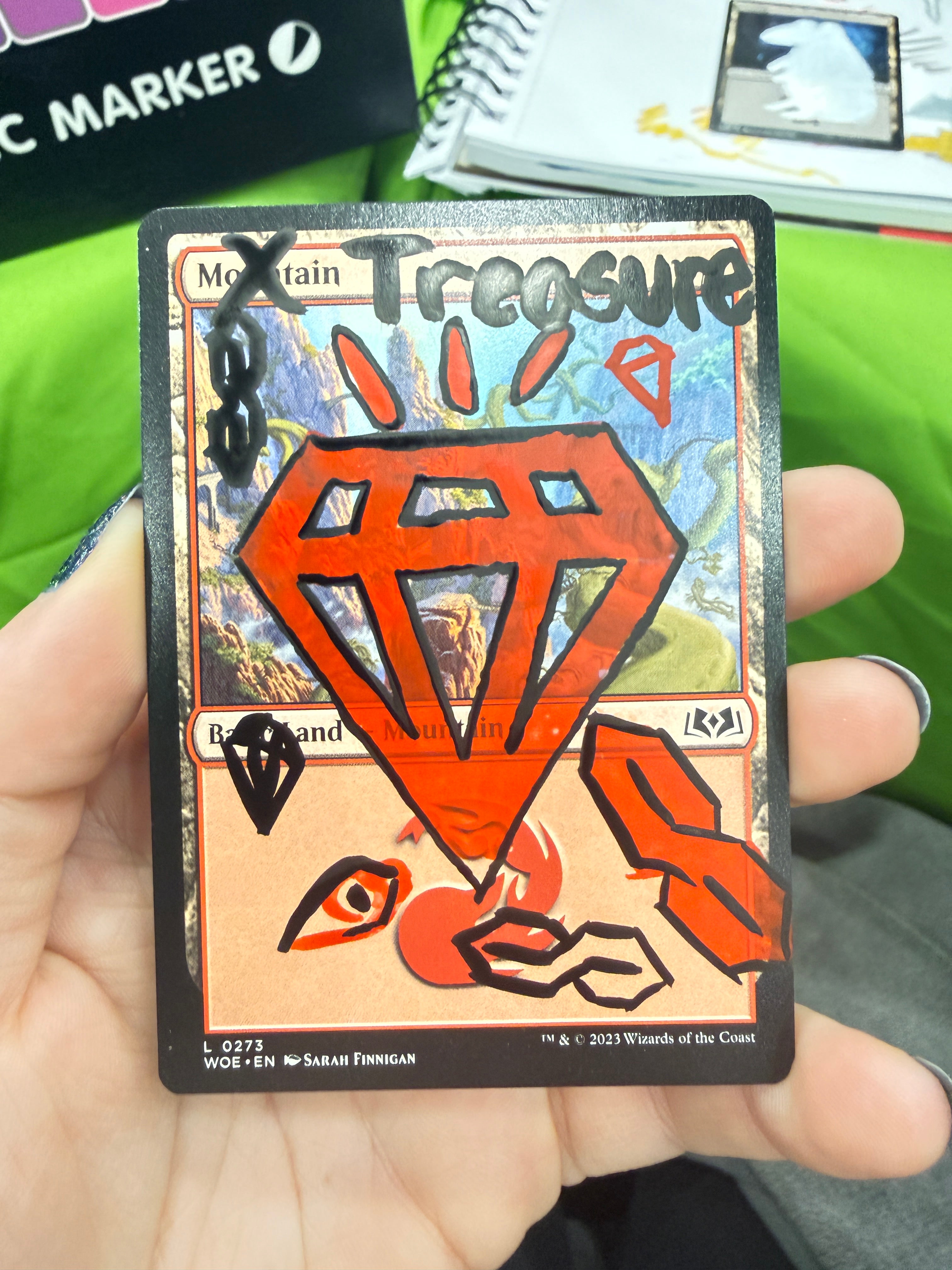 A hand holding a Magic: The Gathering card, specifically a Treasure card with a red gem icon and orange-red artwork, with fingers visible holding it in place.