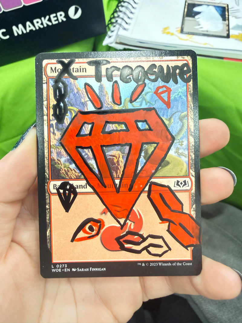 A hand holding a Magic: The Gathering card, specifically a Treasure card with a red gem icon and orange-red artwork, with fingers visible holding it in place.