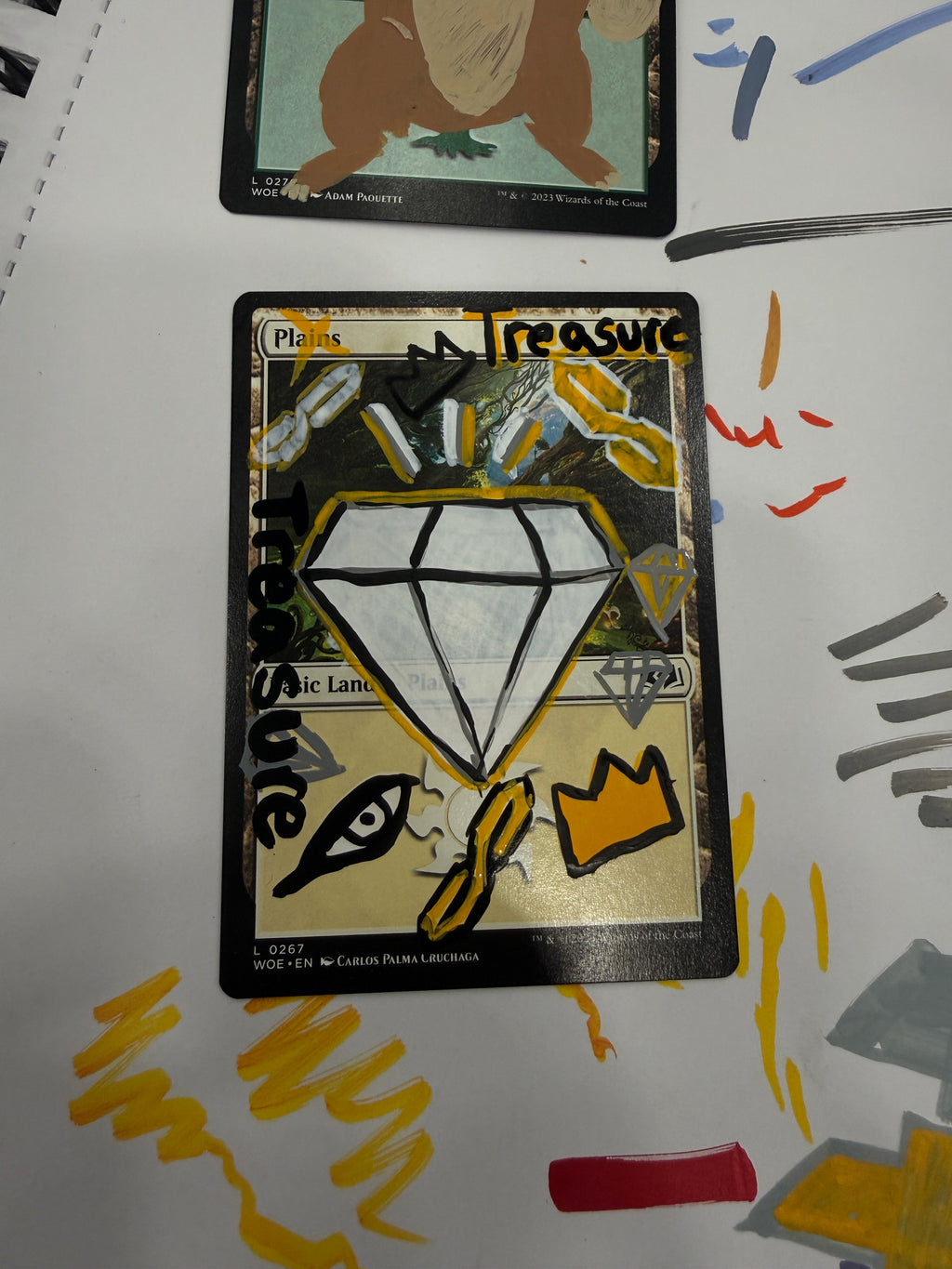 A custom hand-drawn trading card featuring a large white diamond with black outlines, gold background, and the word 'Treasure' written in black. Another card is partially visible on the left, and yellow and gray paint marks are visible on the white surface behind the cards.