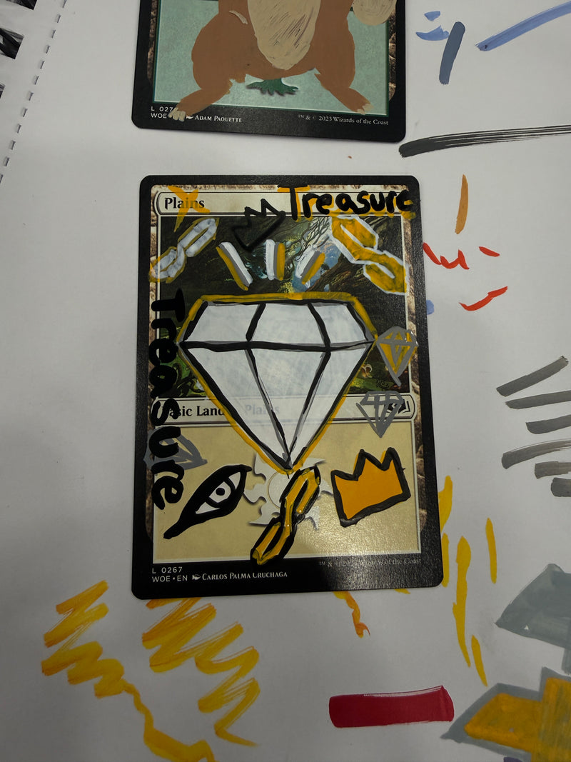 A custom hand-drawn trading card featuring a large white diamond with black outlines, gold background, and the word 'Treasure' written in black. Another card is partially visible on the left, and yellow and gray paint marks are visible on the white surface behind the cards.
